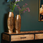 Twee hoge Vaas Liv - Antiek goud | Grote abstracte gezichtsvazen met bloemen staan op een houten tafel met zwart metalen poten en laden, tegen een donkergroene muur met rechts een ingelijst kunstwerk.
