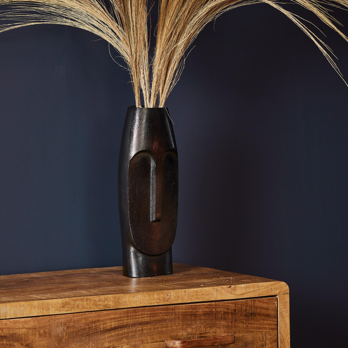 De Vaas Liv - Zwart | Large, met een minimalistisch gezichtsontwerp, houdt hoog gedroogd gras vast op een houten dressoir tegen een donkerblauwe muur.