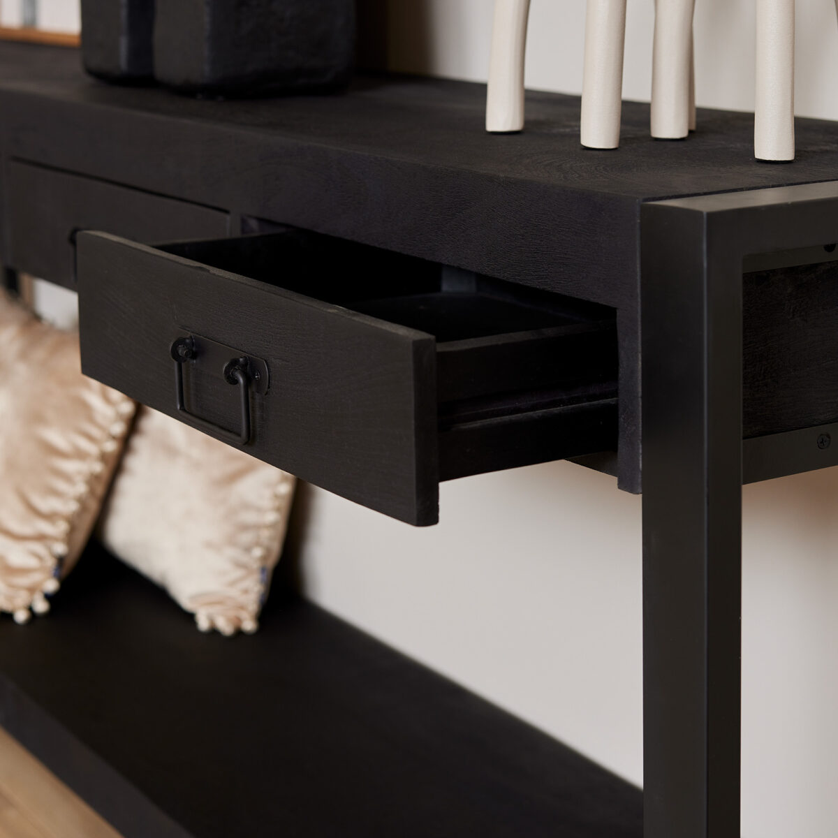 Een close-up van de Sidetable Boaz ZWART | 180 cm toont het moderne zwarte ontwerp met een gedeeltelijk open lade. Decoratieve items liggen bovenop, terwijl zachte beige kussens op de onderste plank liggen.