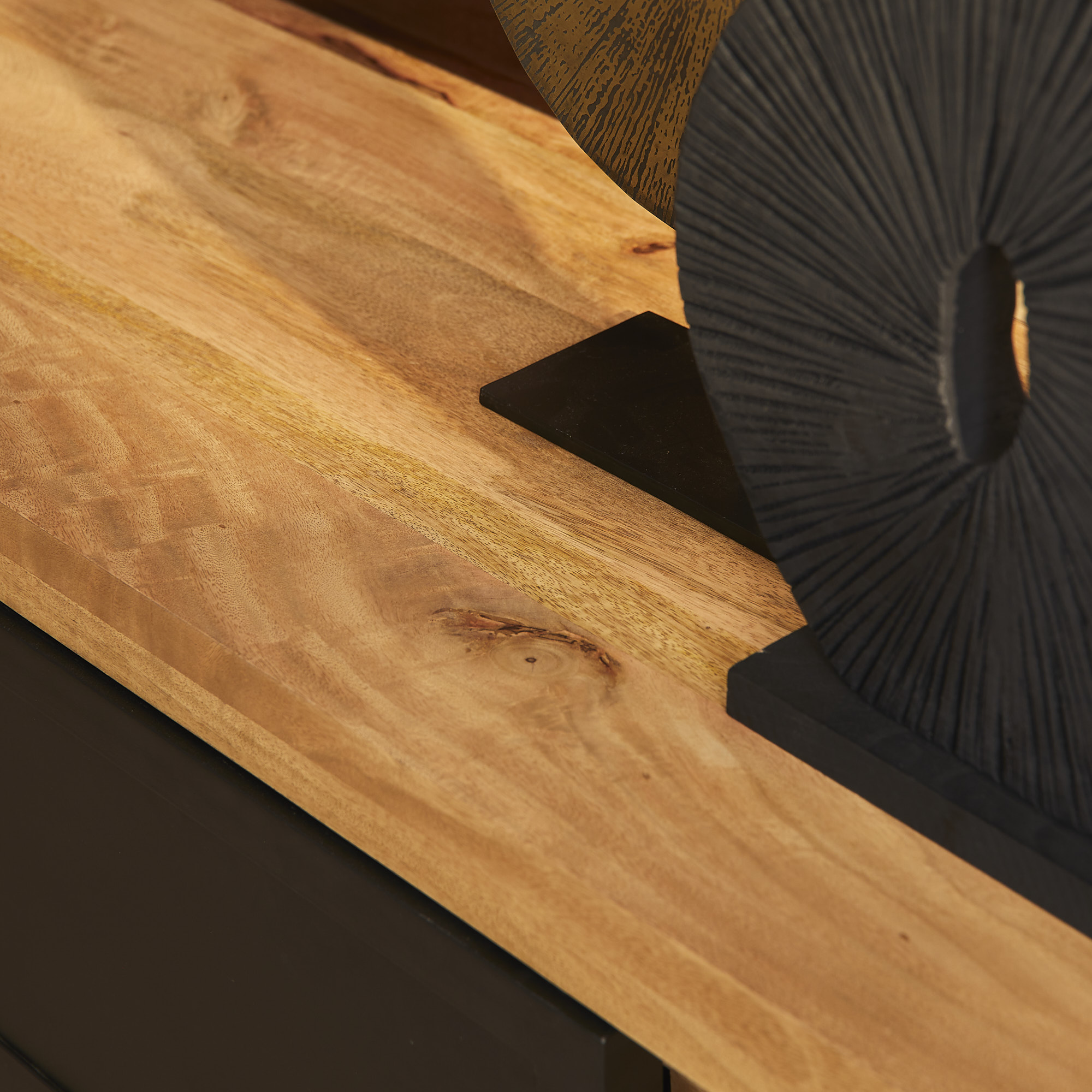 Een close-up van de Nashville Dressoir | 160 cm toont het gladde houten oppervlak met natuurlijke nerf, met een decoratieve zwarte geribbelde ronde sculptuur op een rechthoekige basis in de hoek.