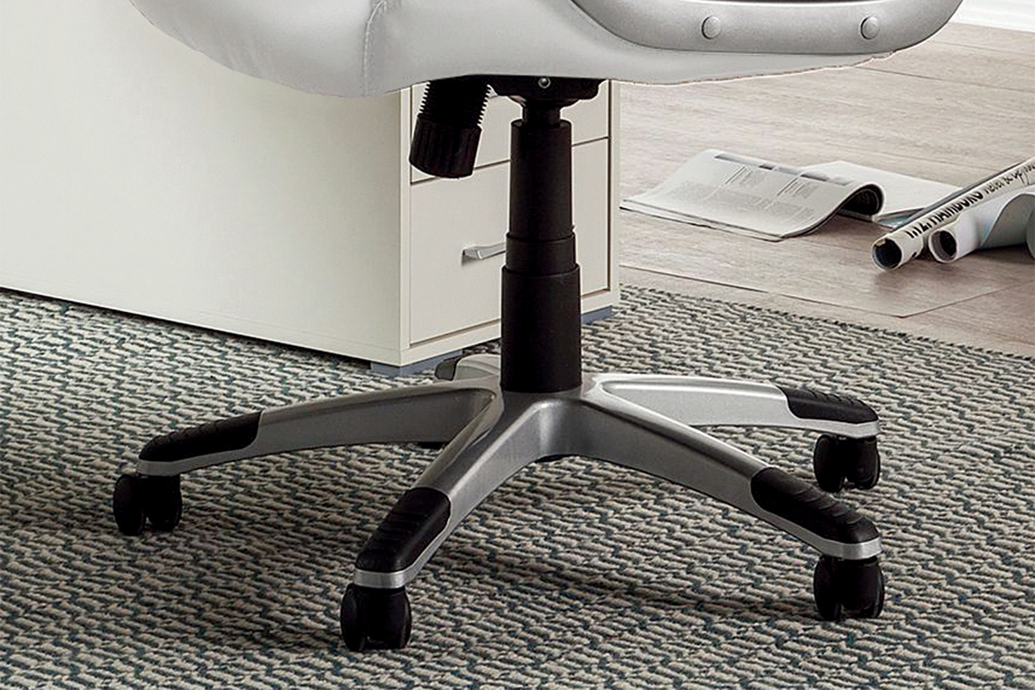 Een close-up van de basis en wielen van de SALE - Bureaustoel 'Kennedy' PU Wit (1c) op een gedessineerd tapijt naast een wit bureau, met opgerolde papieren en een tijdschrift op de houten vloer op de achtergrond.