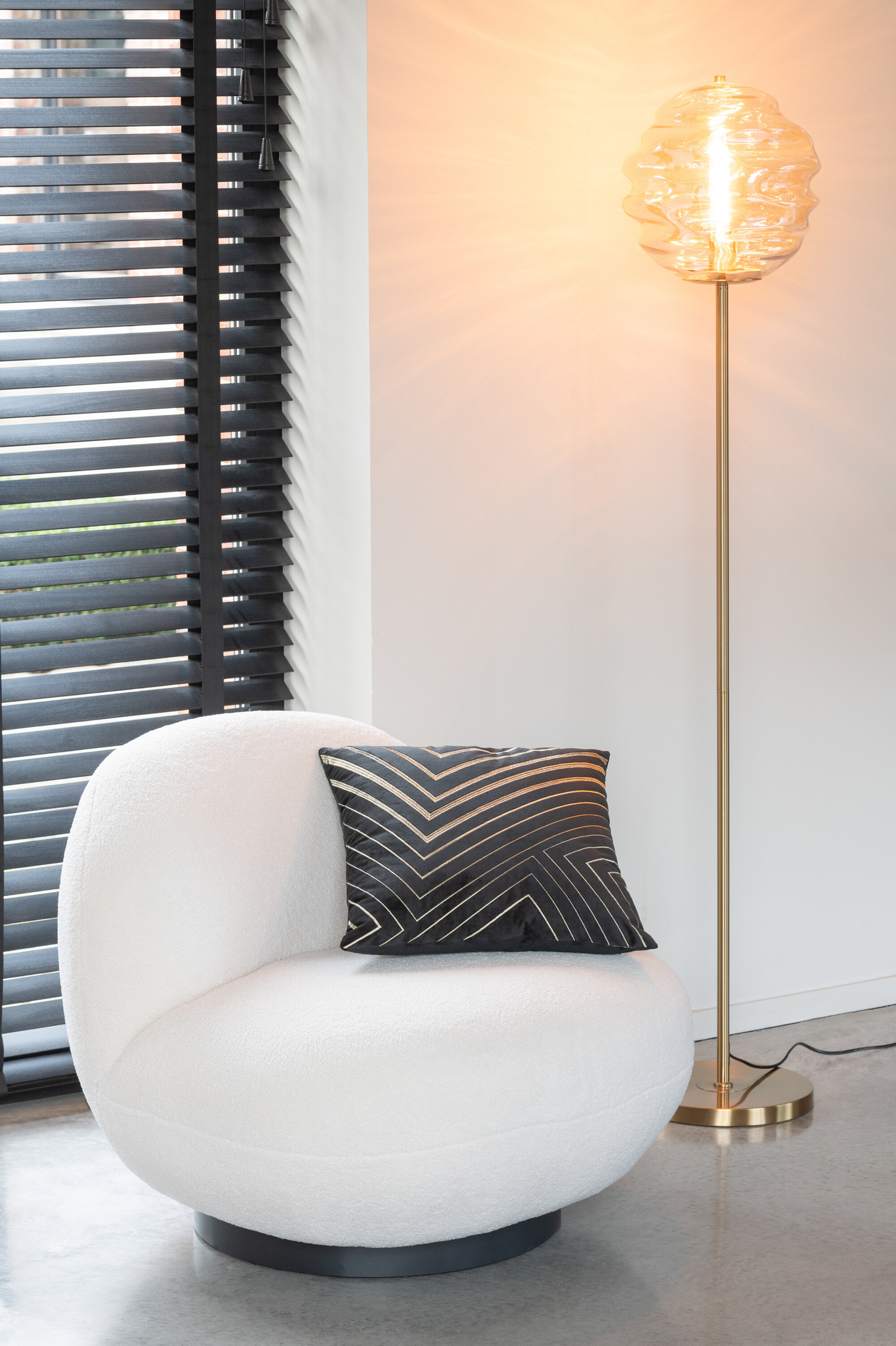 Een moderne leefruimte met de loungestoel Sneeuw Wit/Zwart Comfort en een zwart geometrisch kussen, naast een gouden vloerlamp met een glazen kap met structuur, plus zwarte verticale lamellen voor het raam.