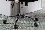 Close-up van de basis van de 'Max' Mesh Zwart bureaustoel met vijf metalen poten op wieltjes, staand op een getextureerd, patroon tapijt naast een wit bureau.