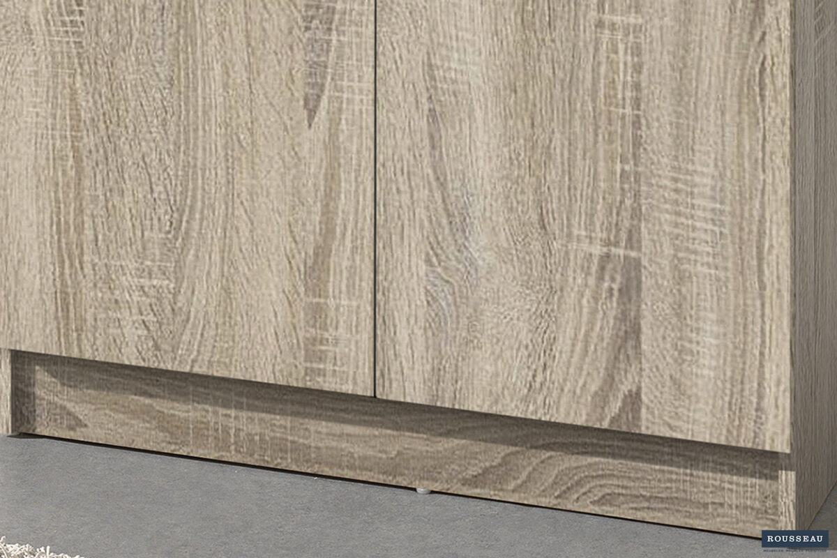 Close-up van het onderste gedeelte van Opbergkast 'Ray' 2 deuren en 4 legplanken Sonoma (1c) met een licht getextureerde houten afwerking, twee deuren en een plint. De vloer is grijs; een klein Rousseau-logo is zichtbaar in de rechterbenedenhoek.