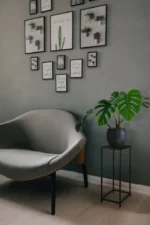 Een moderne, minimalistische kamer met een grijze gebogen stoel en de Mirrors and More Bijzettafel VERONIKA die een monstera in potten vasthoudt, plus ingelijste plantenfoto's en citaten op de grijze muur erboven.