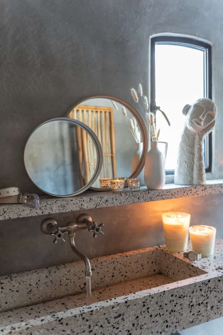 Een terrazzo wastafel met twee brandende kaarsen, een Mirror Round Leather Beige Small, een witte vaas met gedroogde bloemen en een decoratieve sculptuur, tegen een grijze muur en een klein raam.