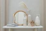 Op een minimaal tafelblad staan keramische vazen, een wandspiegel Round Rattan/Glass Natural Medium, brandende kaarsen, twee huisornamenten, een boek en gedroogd gras tegen lichtbeige muren en gordijnen.