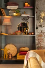 Een gezellige, stijlvolle boekenplank met boeken, kaarsen, een lamp, opgevouwen dekens, kussens, een fles wijn, decoratieve voorwerpen en de Mirror Mona Round Iron/Glass Gold Small als middelpunt, allemaal tegen een bakstenen muur.