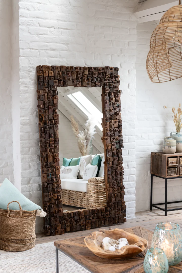 De MIRROR SYMA RECYCLED WOOD/MIRROR NATURAL LARGE met een gestructureerd houten frame leunt tegen een witte bakstenen muur in een gezellige kamer met rieten meubels, zachte kussens, een geweven mand en decoratieve accenten.