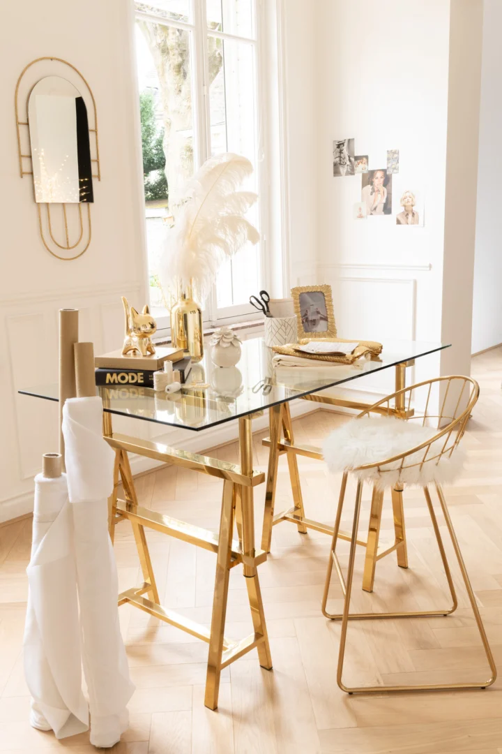 In een lichte, elegante kamer hangt de spiegel Ines Metal Gold boven een glazen bureau met gouden poten, gedecoreerd met boeken en pampagras. Vlakbij staat een gouden stoel met een kussen van wit imitatiebont voor extra stijl.