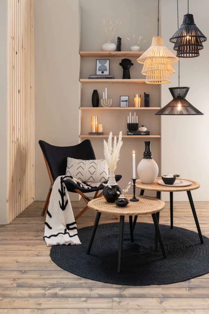 Moderne woonkamer met de Vloerkleed Rond Jute Zwart, houten vloeren, zwarte en rieten meubels, ronde tafels, een dekenstoel met patroon, decoratieve vazen, kaarsen, hanglampen en minimalistische zwart-witte decoraties op planken.