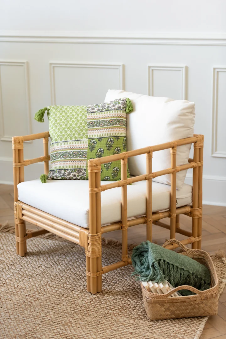 Een rotan fauteuil met witte kussens en een kussen met groen patroon staat op de Vloerkleed Havana Poly Naturel/Wit Klein. Ernaast staat een mand met een groene deken en opgerolde handdoeken in een lichte kamer met wit gelambriseerde muren.