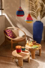 Een gezellige kamer met natuurlijke houten meubels, kleurrijke kaarsen, drankjes, hapjes, hanglampen en een Vloerkleed Rond Gevlochten Zeegras Naturel. Een grote blauwe vaas met pampagras en een kussen met patroon op een geweven stoel voegen charme toe aan de warme, sfeervolle ruimte.