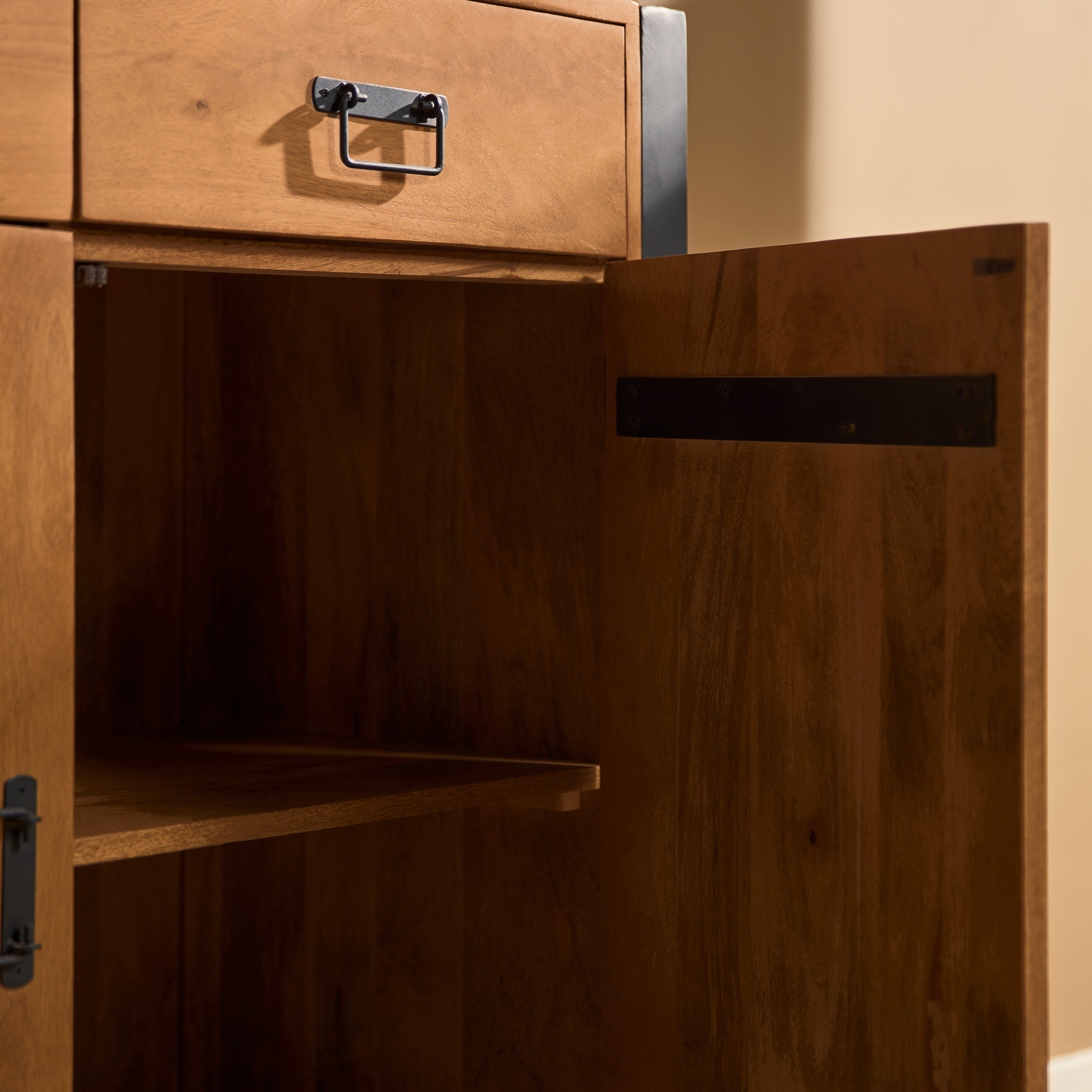 Een close-up van de Vitrinekast Boaz XXL toont een geopende deur die een lege binnenplank onthult. De kast heeft een metalen handvat op de bovenste lade, donker metalen scharnieren op de deur en staat tegen een effen beige muur.