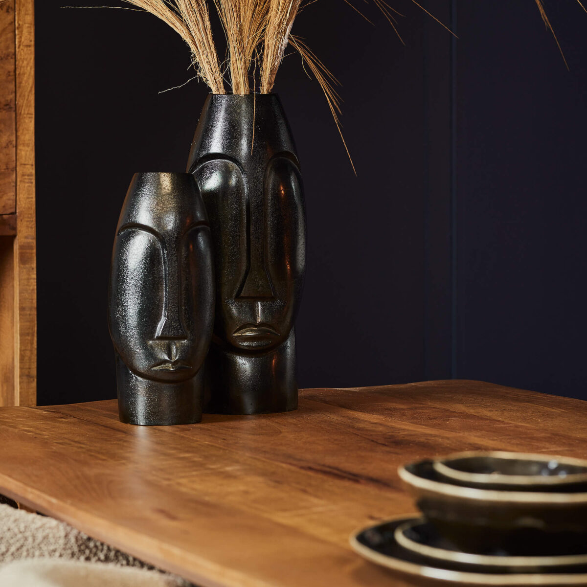 De Vaas Lyra - Zwart | Large, een hoge vaas van zwart keramiek met een abstract gezichtsontwerp, staat met gedroogd gras op een houten tafel naast zwarte en gouden borden tegen een donkere achtergrond.