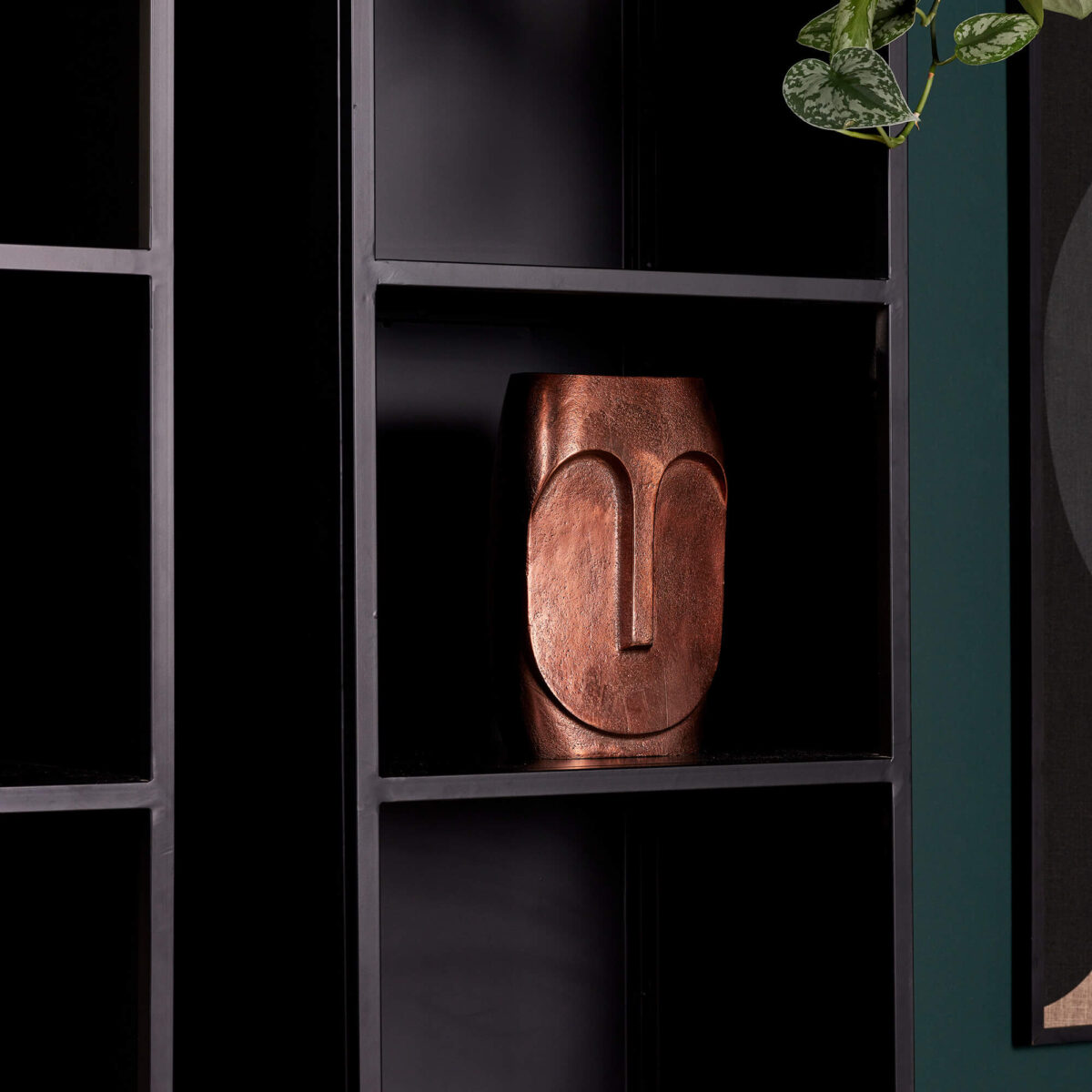 Een bronzen, abstracte gezichtssculptuur staat op de Boekenkast Oakland | 50 cm, een kast in industriële stijl. Rechtsboven zijn een groene bladplant en een donker groenblauwe muur gedeeltelijk zichtbaar.
