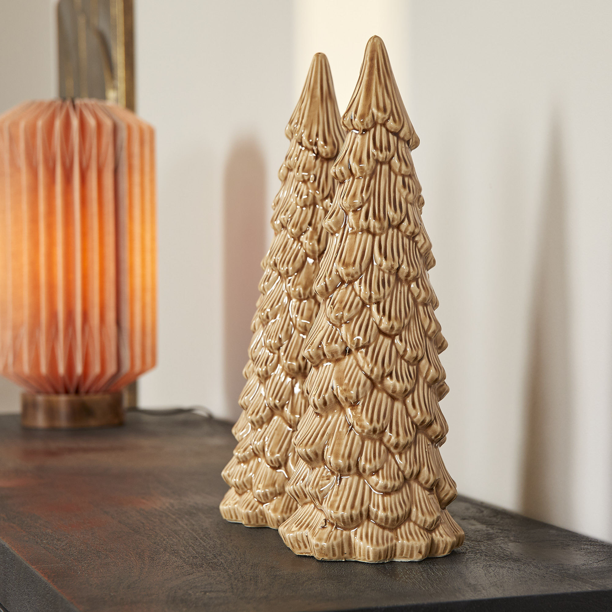 Twee beige keramische dennenboomfiguurtjes zitten op een Dressoir Brussel Black 3 deurs (150 cm). Op de achtergrond straalt een oranje geribbelde lamp een warme gloed uit die de gezellige, moderne binnensetting versterkt.