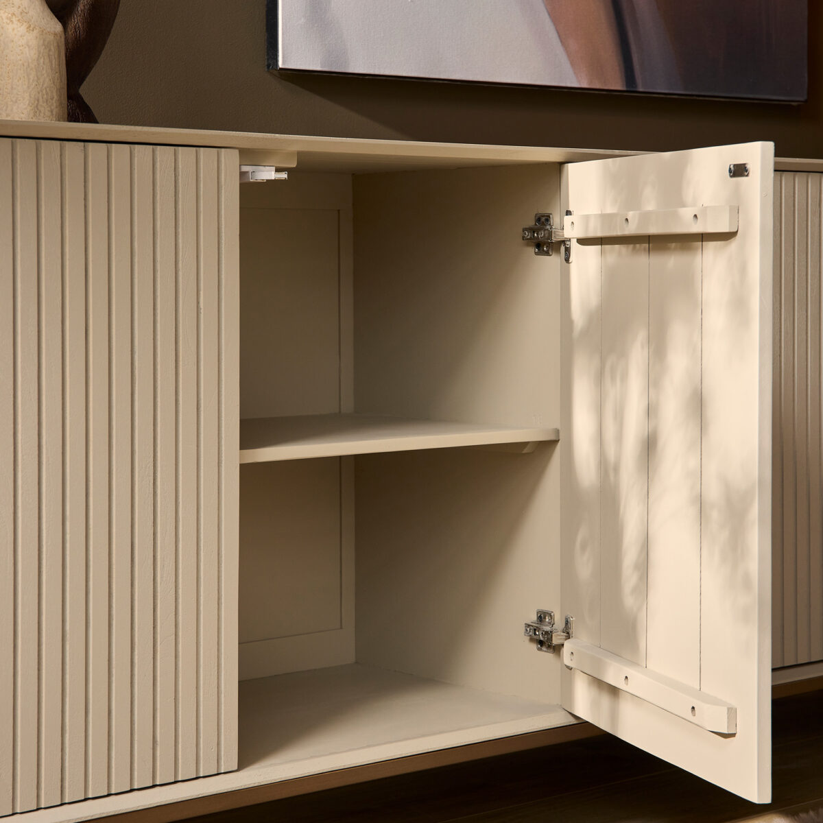 De Dressoir Madison Sand | 210 cm heeft verticale groeven en een open deur die lege planken binnenin onthult, tegen een bruine muur met een deel van een ingelijst kunstwerk erboven.