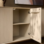 De Dressoir Madison Sand | 210 cm heeft verticale groeven en een open deur die lege planken binnenin onthult, tegen een bruine muur met een deel van een ingelijst kunstwerk erboven.