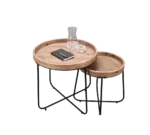 De Mirrors and More Bijzettafel JETTE set bestaat uit twee ronde houten nesttafels met zwarte metalen poten. Op het grotere tafeltje staan een glazen karaf, een waterglas en een gesloten zwart notitieboekje met pen; het kleinere tafeltje is eronder genesteld.