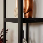 Een close-up van de Boekenkast Boaz Black | 3 lades toont een abstract bronzen gezichtssculptuur en een vaas van crèmekleurig keramiek, met een deel van een decoratieve plant zichtbaar in de linkerbenedenhoek.