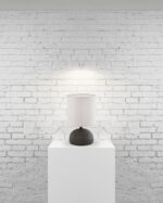 De REALITY Tafellamp LUCI, met een witte kap en donkere voet, is gecentreerd op een witte sokkel tegen een geschilderde bakstenen muur en werpt zacht licht naar boven.