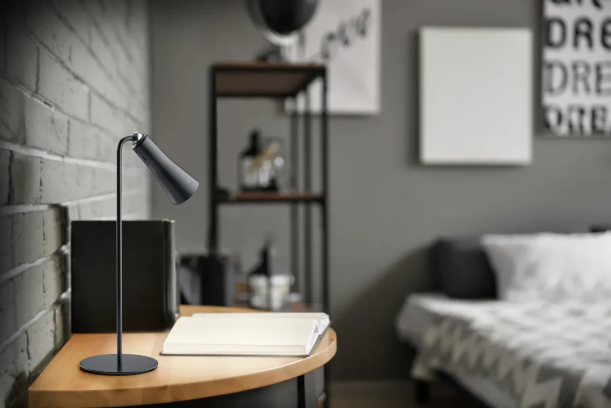 De Reality Tafellamp i zit op een houten bureau met een open notitieblok en pen, tegen een grijze bakstenen muur in een stijlvolle, minimalistische slaapkamer met vage planken en een bed op de achtergrond.