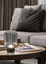 De REALITY Tafellamp LORD, met een geribbelde glazen kap, staat op een houten salontafel bij een open tijdschrift voor een beige bank met bruine en grijze kussens en kamerhoge gordijnen.
