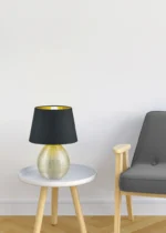 Een ronde witte bijzettafel met de REALITY Tafellamp LUXOR, met een gestructureerd gouden onderstel en donkere kap, staat naast een lichthouten fauteuil met donkergrijze bekleding op een visgraat houten vloer tegen een effen witte muur.