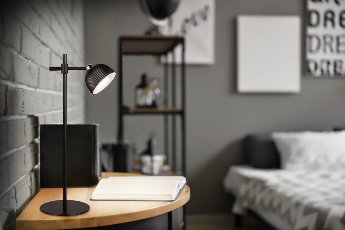 De REALITY Tafellamp MAXIMA, een moderne zwarte bureaulamp, staat op een houten bureau met een open notitieblok in een slaapkamer met een grijs thema, een bed, decoratieve planken en abstracte muurkunst op de achtergrond.