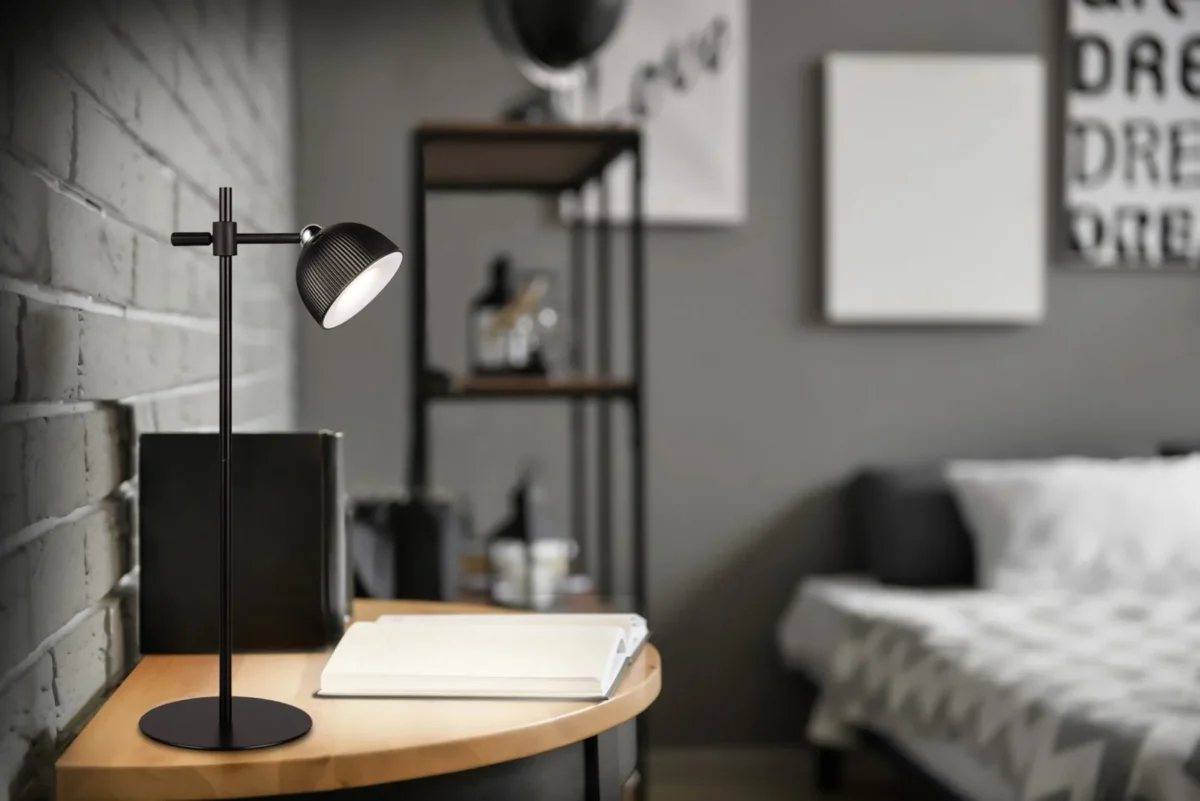 De REALITY Tafellamp MAXIMA, een moderne zwarte bureaulamp, staat op een houten bureau met een open notitieblok in een slaapkamer met een grijs thema, een bed, decoratieve planken en abstracte muurkunst op de achtergrond.