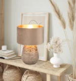 Een decoratieve tafel met de Reality Tafellamp Kiran, ingelijste kunstwerken, boeken, een witte vaas met gedroogde bloemen en geweven opbergmanden eronder, allemaal tegen een lichtgekleurde muur.