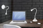 Een modern bureau met een laptop, draadloze muis, smartphone en twee REALITY Tafellamp VIPER lampen - één op het bureau en één aan de muur - tegen een achtergrond van donkere bakstenen muren.