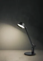 De TRIO Tafellamp Ava L, een moderne zwarte bureaulamp met een verstelbare arm, verlicht een effen beige oppervlak tegen een donkere achtergrond met textuur.