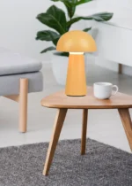 De REALITY Tafellamp LENNON, een moderne gele paddenstoelvormige lamp, staat op een houten salontafel naast een witte mok in een lichte woonkamer met een grijze bank, groene plant en grijs vloerkleed.