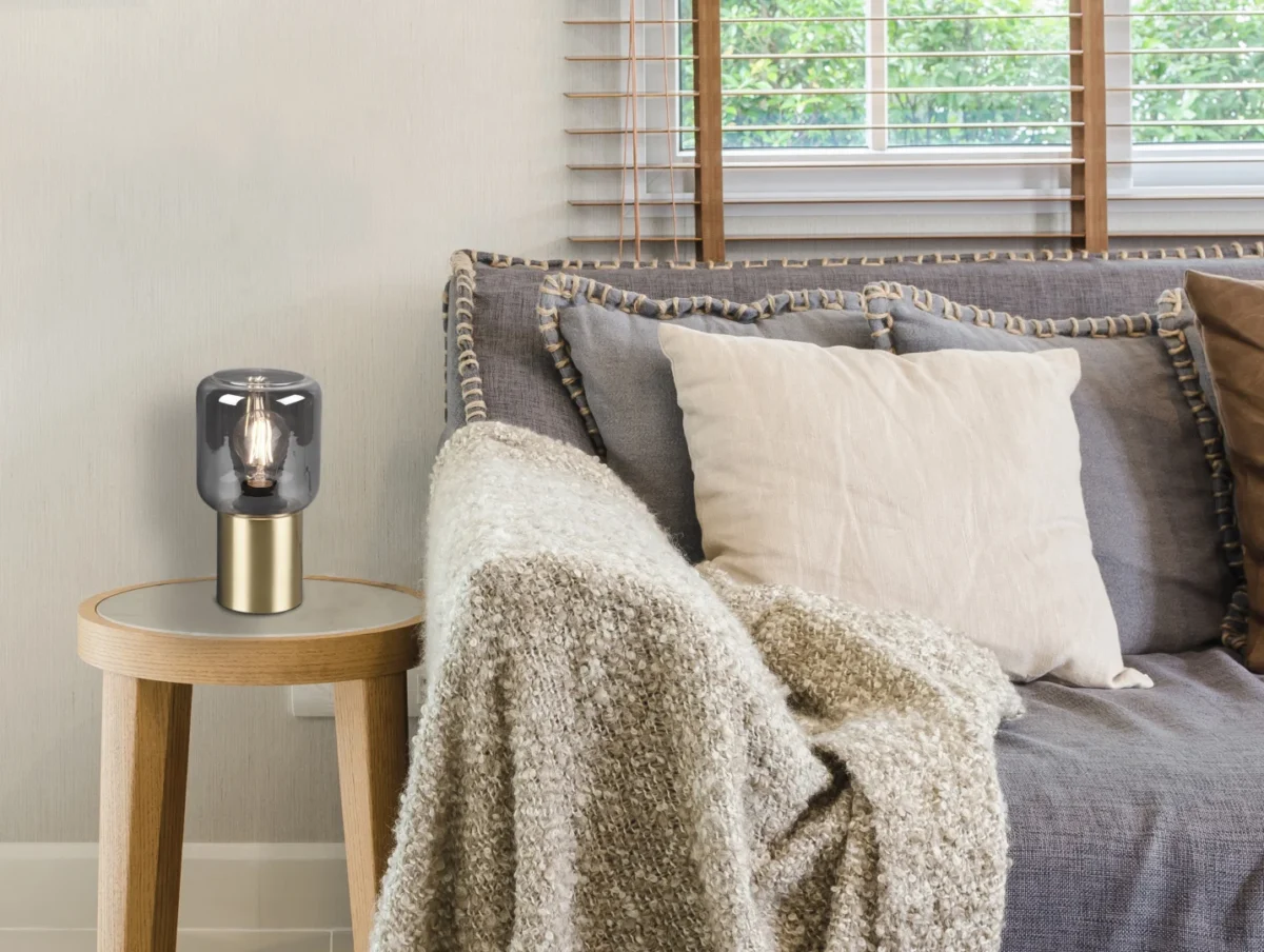 Een gezellig huiskamertafereel met een grijze bank, beige en grijze kussens, een structuurdeken en een houten bijzettafeltje met de REALITY Tafellamp NICO bij een raam met houten jaloezieën en groen buiten.