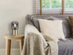 Een gezellig huiskamertafereel met een grijze bank, beige en grijze kussens, een structuurdeken en een houten bijzettafeltje met de REALITY Tafellamp NICO bij een raam met houten jaloezieën en groen buiten.