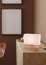 De Reality Tafellamp Woody, met een rechthoekige witte kap en houten voet, staat op een lichthouten kast onder twee lege frames op een bruine muur, met een bruinleren stoel gedeeltelijk in het zicht.