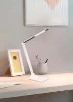 De TRIO Tafellamp Travis, een moderne witte bureaulamp, staat op een licht houten bureau naast een fotolijst, een ronde pennenpot en een open notitieblok. Minimalistische kunst siert de lichtgrijze muur erachter.