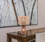 De REALITY Tafellamp STRAW staat op een houten eettafel met twee witte stoelen. Achter de tafel hangt een groot wandkunstwerk met abstracte boomtakken in neutrale tinten.