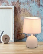 De REALITY Tafellamp MALU, een kleine witte tafellamp met een stoffen kap, staat op een houten bureau naast een leeg canvas, een ronde klok en een pennenbeker, tegen een rustieke muur met textuur.