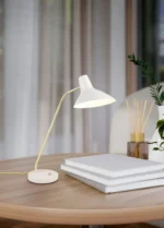 De TRIO Tafellamp Traveller, een moderne witte lamp, wordt aangezet en verlicht een ronde houten tafel met drie gestapelde boeken, een plant en een rietverdamper in een gezellige, zacht verlichte kamer.