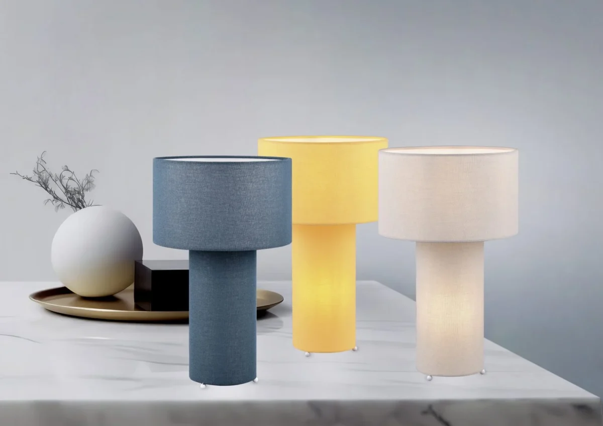 Drie TRIO tafellampen Bale - blauw, geel en crème - worden getoond op een wit marmeren oppervlak. Een decoratief dienblad met een rond wit object en gedroogde takken staat op de achtergrond.