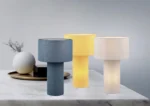 Drie TRIO tafellampen Bale - blauw, geel en crème - worden getoond op een wit marmeren oppervlak. Een decoratief dienblad met een rond wit object en gedroogde takken staat op de achtergrond.