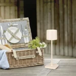 Een rieten picknickmand met borden en bestek staat op een houten oppervlak naast het Reality Tafellamp Fernandez, tegen een rustieke houten achtergrond.
