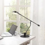 De TRIO Manduro tafellamp, een open laptop en een witte koptelefoon liggen op een wit bureau bij een helder raam met transparante gordijnen, waardoor de ruimte wordt gevuld met natuurlijk licht en uitzicht op het groen buiten.