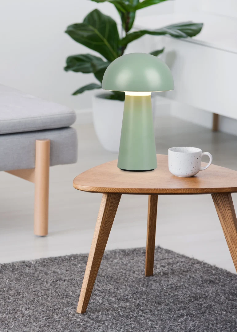 De REALITY Tafellamp LENNON, een kleine groene paddenstoelvormige lamp, staat op een houten tafel naast een witte mok in een moderne woonkamer met een grijze bank, groene plant en grijs vloerkleed.