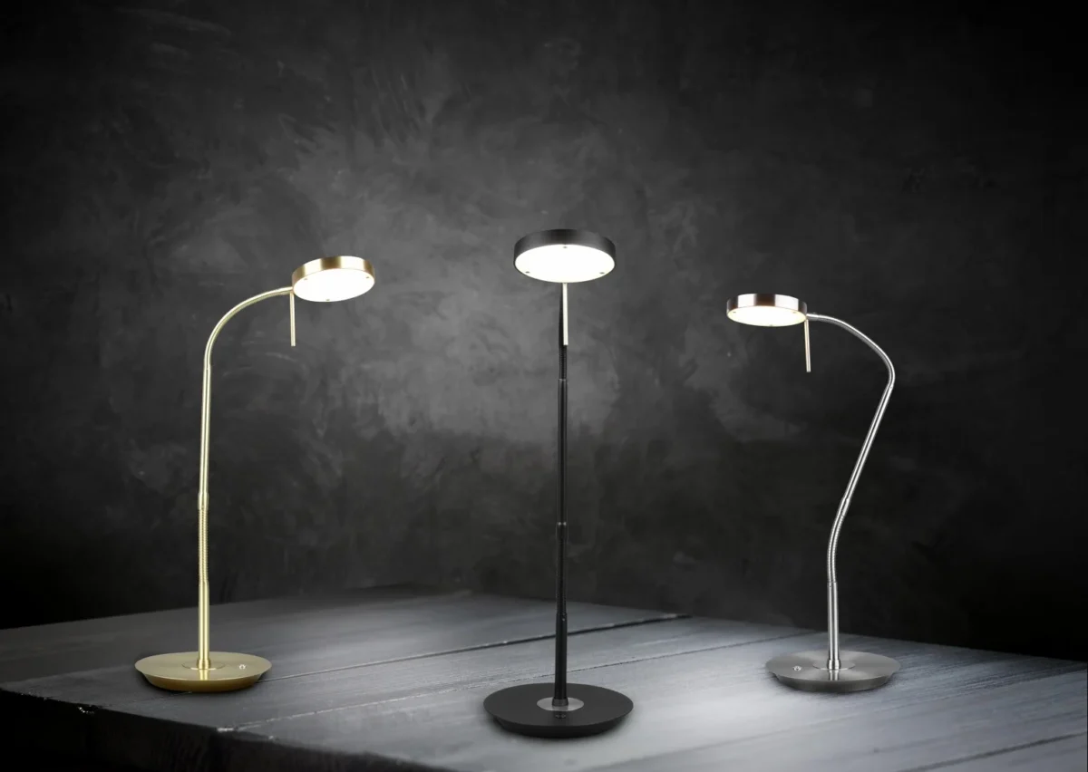 Drie TRIO Tafellamp Monza bureaulampen in goud, zwart en zilver worden verlicht op een donker houten oppervlak tegen een gestructureerde achtergrond, elk met een ronde voet, slanke steel en ronde kop.