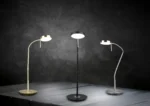 Drie TRIO Tafellamp Monza bureaulampen in goud, zwart en zilver worden verlicht op een donker houten oppervlak tegen een gestructureerde achtergrond, elk met een ronde voet, slanke steel en ronde kop.