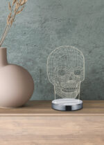 De REALITY Tafellamp SKULL, een draadvormige lamp in de vorm van een schedel met een metalen voet, is geplaatst op een houten oppervlak naast een matbeige vaas met gedroogde takken tegen een groene muur met textuur.